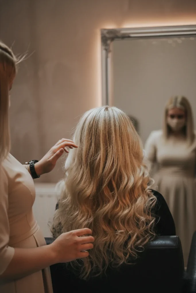 A professional hairstylist in a beige dress styling a client's long, wavy hair with blonde highlights and balayage at Seoyeon Beauty Salon in Al Ain.